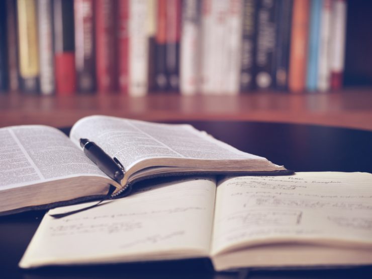 In the forefront, on a table an opened notebook and an opened book, with a pencil laying in the middle of it. In the background books on a bookshelf.