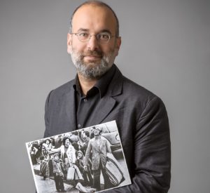 Picture of Felipe Estrada, holding a picture of his family as they arrive in Sweden in 1974. Felipe has greyish beard, wears round glasses and is dressed in a grey suit and dark-grey shirt.