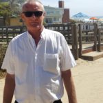 Professor John Pratt standing in front of a fence and some houses, close to a beach. Professor Pratt wears sunglasses and a white shirt.