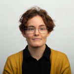 Portrait picture of PhD-student Aino Jauhiainen, Helsinki University. Aino has short, brown, curly hair and wear glasses. She is dressed in a black shirt and yellow cardigan.