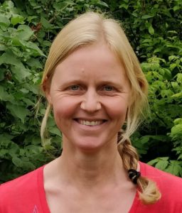Photo of Mie Birk Haller. She is standing outside, green leaves in the background, dressed in a red t-shirt. She has blond hair in a braid and is smiling.