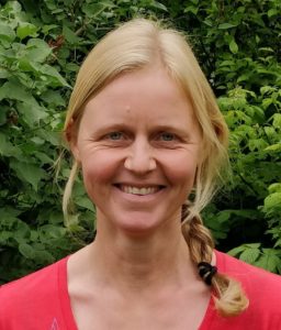 Photo of Mie Birk Haller. She is standing outside, green leaves in the background, dressed in a red t-shirt. She has blond hair in a braid and is smiling.