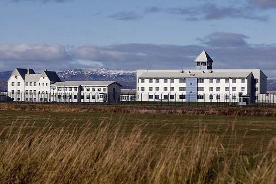 A photo of Litla-Hraun, Iceland´s largest prison, from a distance. A large, white, long building surrounded by wire fences and green fields, against a mountainous backdrop. 