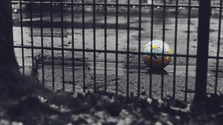 Photo of a yellow football lying on wet asphalt behind a fence. 
